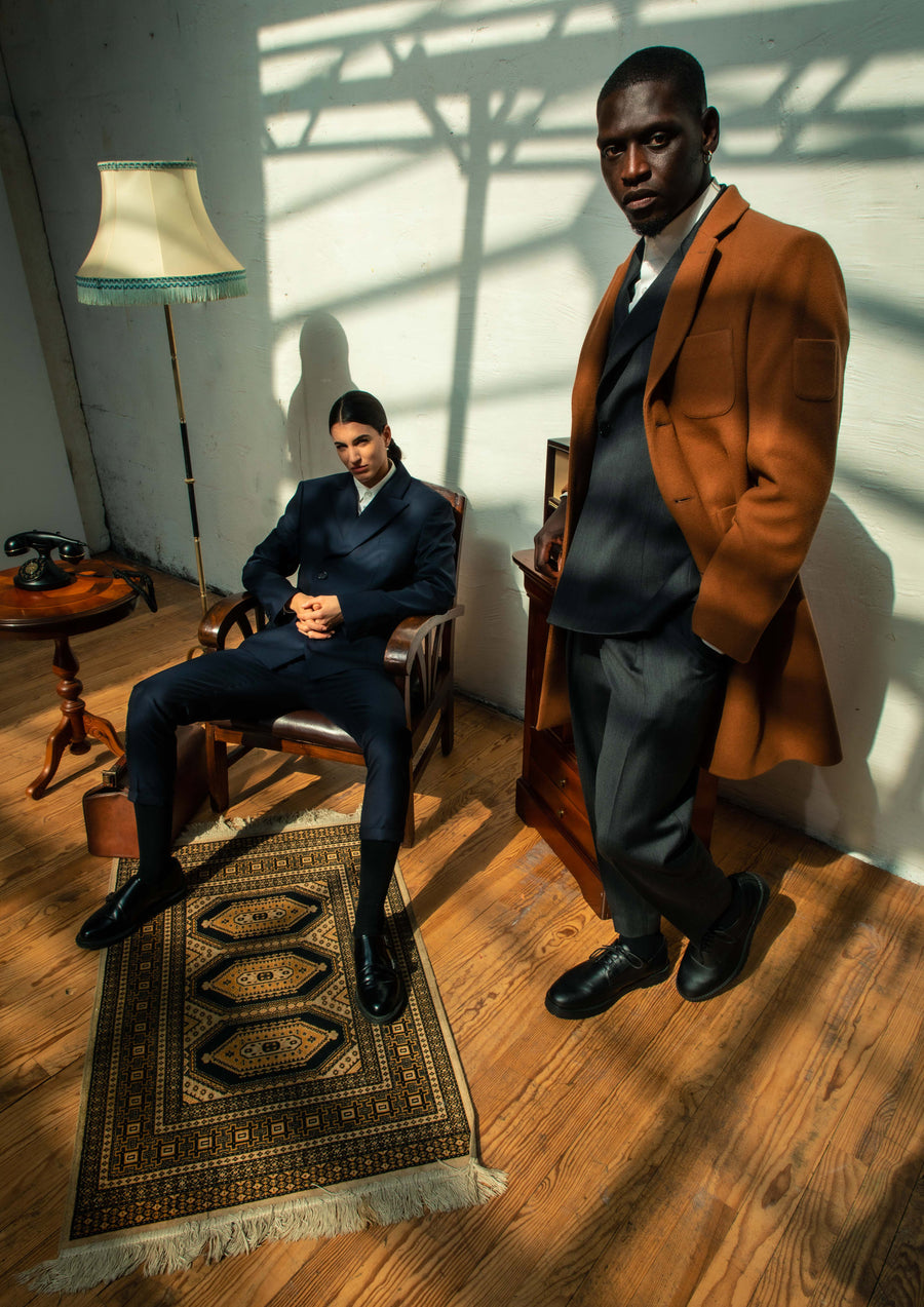 Homme debout en manteau tabac sur un costume gris, un second assis en costume bleu marine, dans un salon ensoleillé